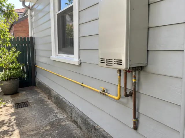 Yellow gas pipe and fittings on an exterior weatherboard wall beside a window in Petersham