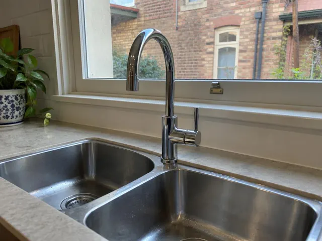 Chrome kitchen mixer tap over a stainless steel double sink beside a window in a brick home interior in Newtown